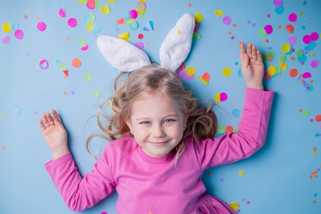 Piccola ragazza bionda sveglia nelle orecchie del coniglietto di Pasqua in vestito rosa su sfondo blu.