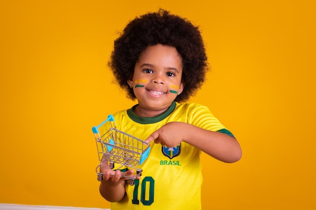 Piccola ragazza brasiliana cheerleader in abito da calcio che tiene un mini carrello in mano Concetto di shopping e promozione della Coppa del mondo