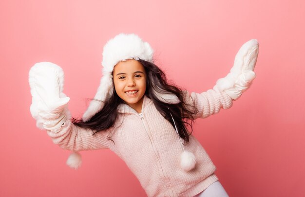 Piccola ragazza bruna con i capelli lunghi in un cappello invernale e guanti su uno sfondo rosa