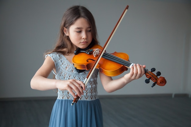 Piccola ragazza bruna in un bellissimo vestito che suona il violino al chiuso Concetto di strumenti musicali