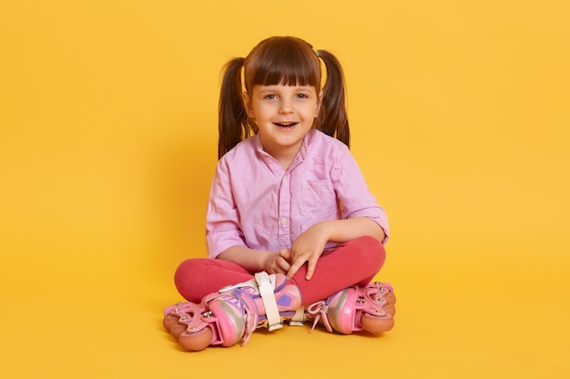 Piccola ragazza carina affascinante con pattini a rotelle seduto sul pavimento con le gambe incrociate