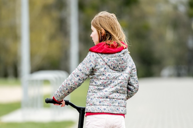 Piccola ragazza carina che guida uno scooter su un sentiero nel parco