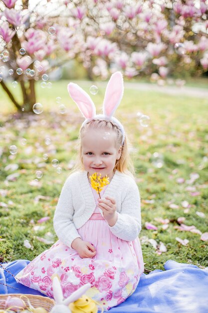 Piccola ragazza carina che si siede sull'erba vicino alla magnolia. Una ragazza vestita da coniglietto pasquale tiene in mano un fiore e un uovo. Primavera.