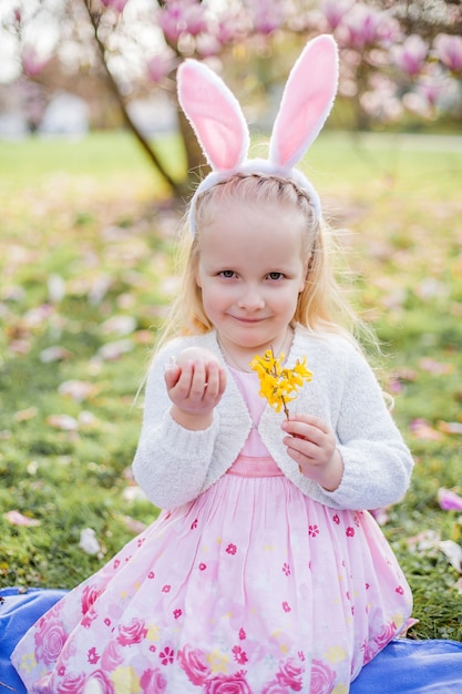 Piccola ragazza carina che si siede sull'erba vicino alla magnolia. Una ragazza vestita da coniglietto pasquale tiene in mano un fiore e un uovo. Primavera.