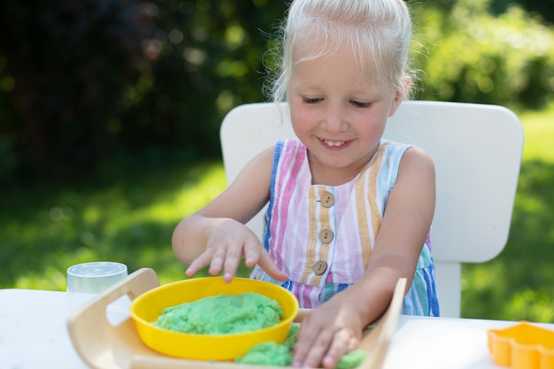 Piccola ragazza carina con i capelli biondi che gioca con la sabbia cinetica colorata all'aperto nel cortile di casa in una giornata estiva. Hobby, attività per il tempo libero, artigianato, modellismo e concetto di hobby creativo