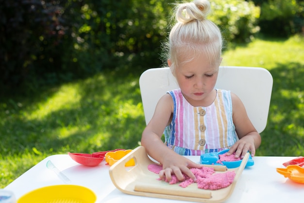 Piccola ragazza carina con i capelli biondi che gioca con la sabbia cinetica colorata all'aperto nel cortile di casa in una giornata estiva. Hobby, attività per il tempo libero, artigianato, modellismo e concetto di hobby creativo