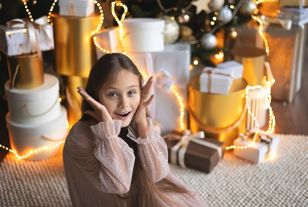 Piccola ragazza carina con i capelli lunghi in abito seduto sul pavimento e apre una scatola con un regalo per lo sfondo albero di Natale Xmas concetto di vacanza