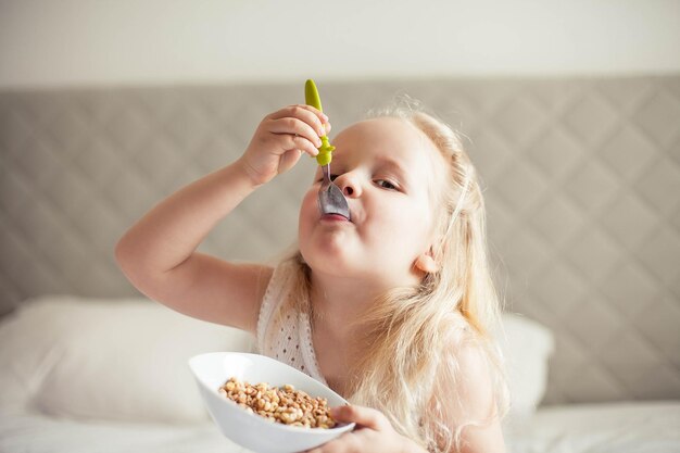 Piccola ragazza carina dai capelli bianchi mangia muesli sul letto nella sua stanza luminosa Cibo sano Comfort domestico