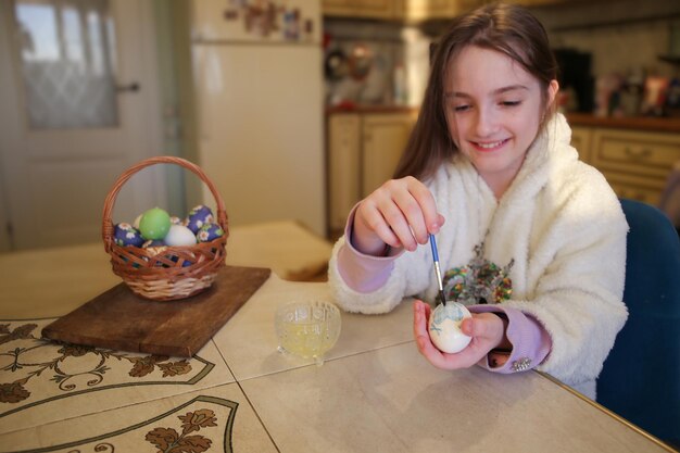 Piccola ragazza carina felice che decora le uova di Pasqua usando pennello e vernice in cucina