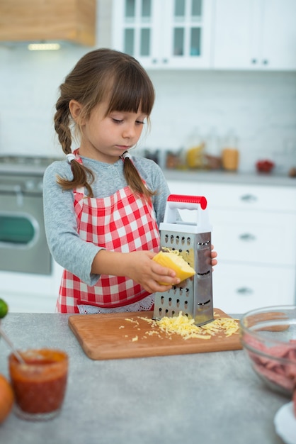 Piccola ragazza carina sorridente con le trecce e in un grembiule da cucina a scacchi strofina il formaggio su una grattugia, aiuta a cucinare la pizza in cucina.