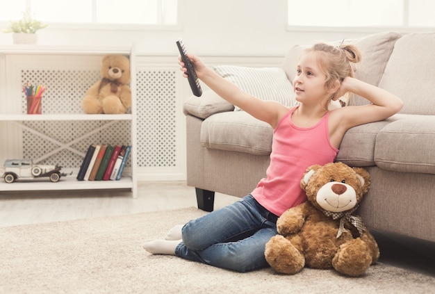 Piccola ragazza casuale che guarda la tv a casa. Bambina seduta sul tappeto del pavimento con il suo amico giocattolo orsacchiotto, che tiene in mano il telecomando e cambia canale, copia spazio