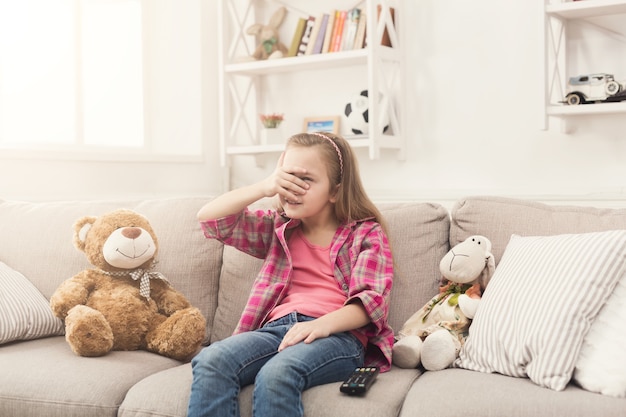 Piccola ragazza casuale spaventata che guarda la tv Bambina spaventata seduta sul divano con gli occhi chiusi, a casa da sola, guardando film spaventosi proibiti con i suoi amici giocattolo orsacchiotto e pecora, copia spazio