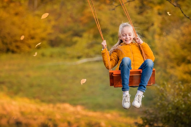 Piccola ragazza caucasica bionda felice che sorride e che cavalca un'altalena di corda in autunno nel parco