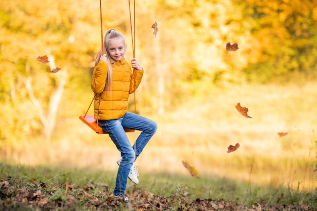 Piccola ragazza caucasica bionda felice che sorride e che cavalca un'altalena di corda in autunno nel parco