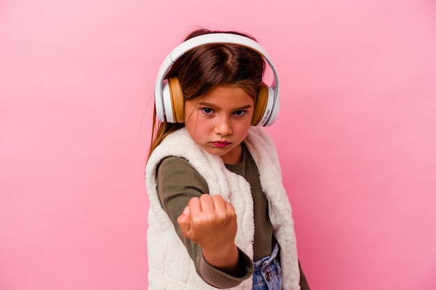 Piccola ragazza caucasica che ascolta musica isolata su sfondo rosa che mostra pugno alla telecamera, espressione facciale aggressiva.