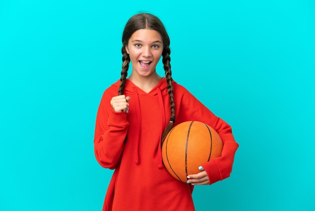 Piccola ragazza caucasica che gioca a basket isolata su sfondo blu che celebra una vittoria in posizione di vincitore