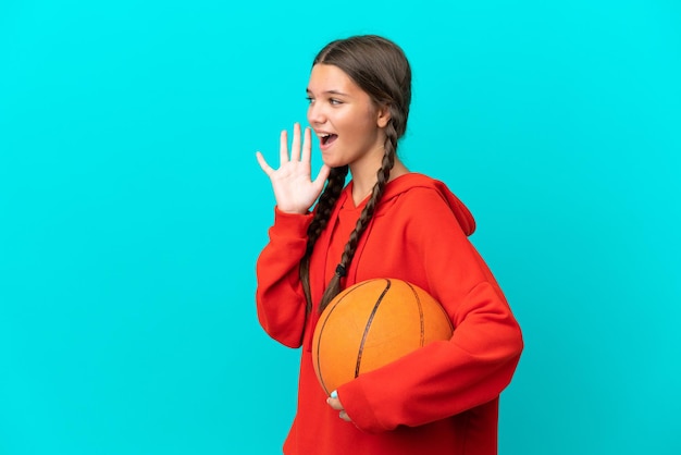 Piccola ragazza caucasica che gioca a basket isolata su sfondo blu che grida con la bocca spalancata di lato