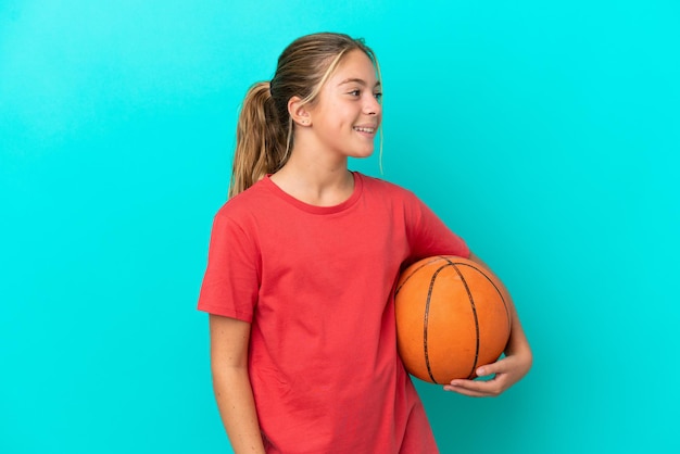 Piccola ragazza caucasica che gioca a basket isolata su sfondo blu che guarda al lato e sorridente