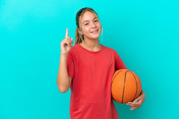 Piccola ragazza caucasica che gioca a basket isolata su sfondo blu che mostra e alza un dito in segno di meglio