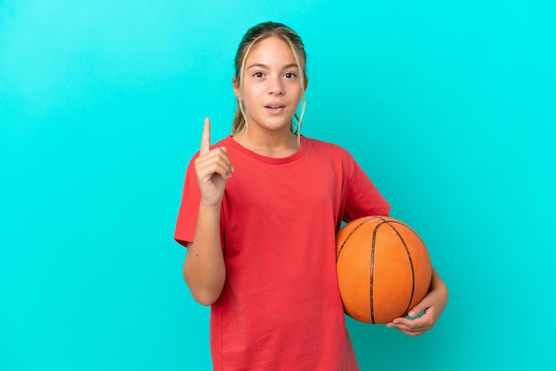 Piccola ragazza caucasica che gioca a basket isolata su sfondo blu con l'intenzione di realizzare la soluzione sollevando un dito