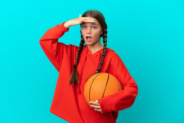 Piccola ragazza caucasica che gioca a basket isolata su sfondo blu facendo un gesto a sorpresa mentre guarda di lato