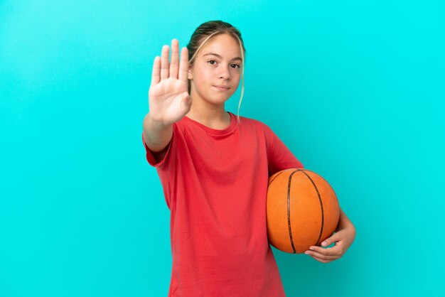 Piccola ragazza caucasica che gioca a basket isolata su sfondo blu facendo un gesto di arresto