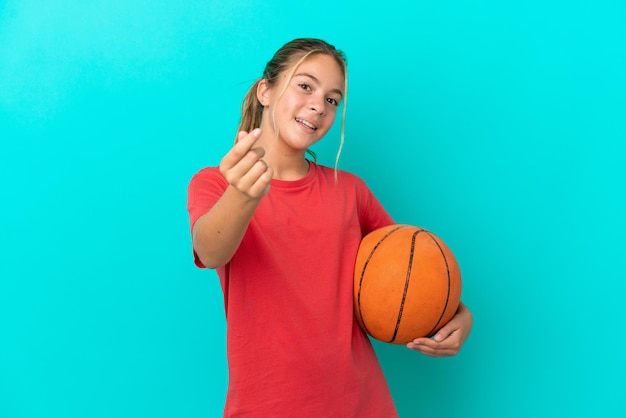 Piccola ragazza caucasica che gioca a basket isolata su sfondo blu facendo un gesto di denaro