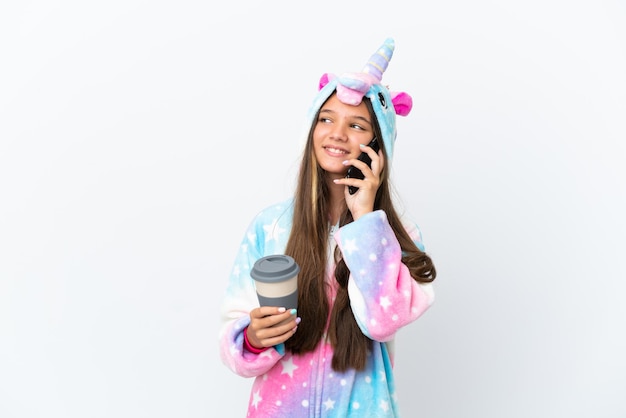 Piccola ragazza caucasica che indossa un pigiama unicorno isolato su sfondo bianco che tiene il caffè da portare via e un cellulare