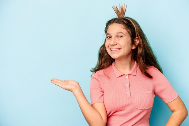 Piccola ragazza caucasica che indossa una corona principessa isolata su sfondo blu che mostra uno spazio di copia su un palmo e tiene un'altra mano sulla vita.