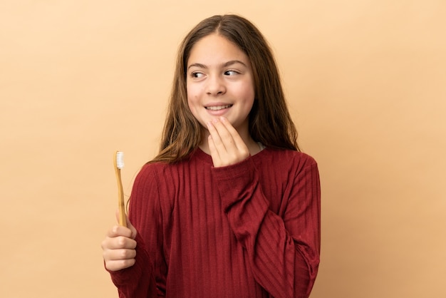 Piccola ragazza caucasica che si lava i denti isolati su sfondo beige alzando lo sguardo mentre sorride