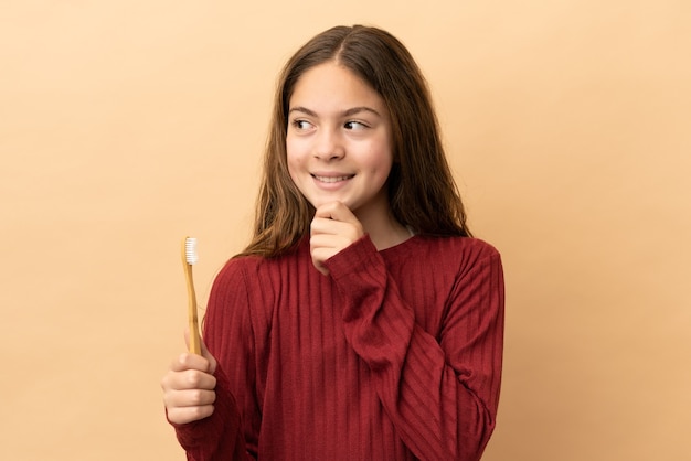 Piccola ragazza caucasica che si lava i denti isolati su sfondo beige e alza lo sguardo