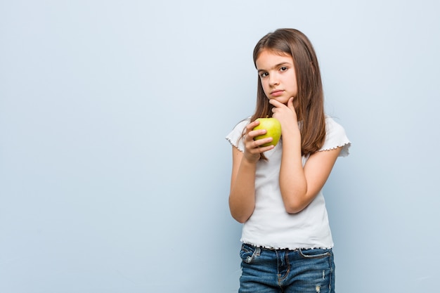 Piccola ragazza caucasica che tiene mela verde lateralmente con espressione dubbiosa e scettica.