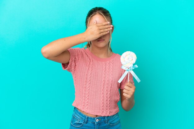 Piccola ragazza caucasica che tiene un lecca-lecca isolato su sfondo blu che copre gli occhi con le mani. Non voglio vedere qualcosa