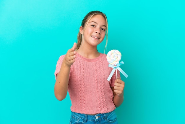 Piccola ragazza caucasica che tiene un lecca-lecca isolato su sfondo blu che stringe la mano per chiudere un buon affare