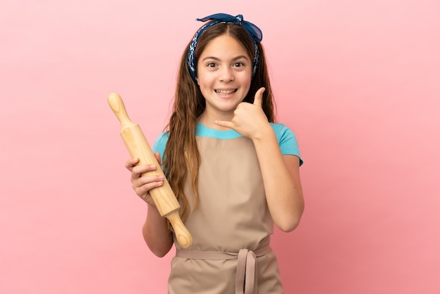 Piccola ragazza caucasica che tiene un mattarello isolato su sfondo rosa che fa il gesto del telefono. Richiamami segno