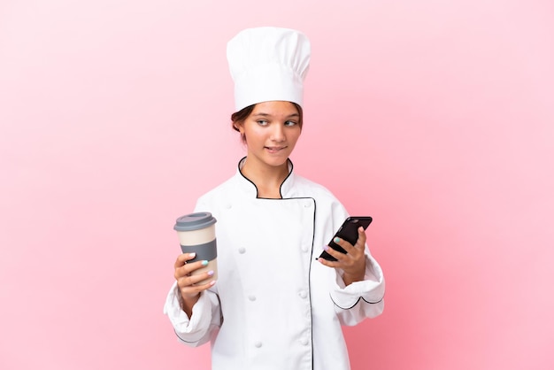 Piccola ragazza caucasica chef isolata su sfondo rosa che tiene il caffè da portare via e un cellulare mentre pensa a qualcosa