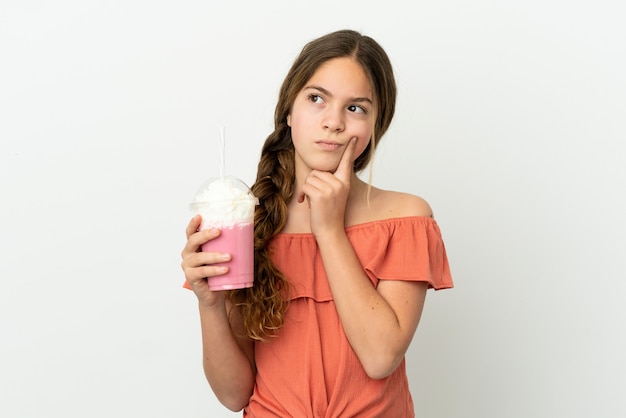 Piccola ragazza caucasica con frullato di fragole isolato su sfondo bianco che ha dubbi mentre guarda in alto