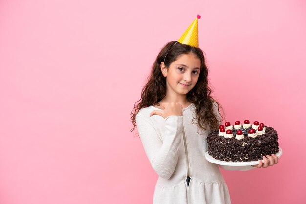 Piccola ragazza caucasica con torta di compleanno isolata su sfondo rosa che punta di lato per presentare un prodotto