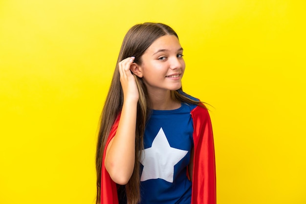 Piccola ragazza caucasica del supereroe isolata su fondo giallo che ascolta qualcosa mettendo la mano sull'orecchio