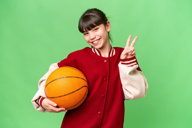 Piccola ragazza caucasica giocare a basket su sfondo isolato sorridente e mostrando segno di vittoria