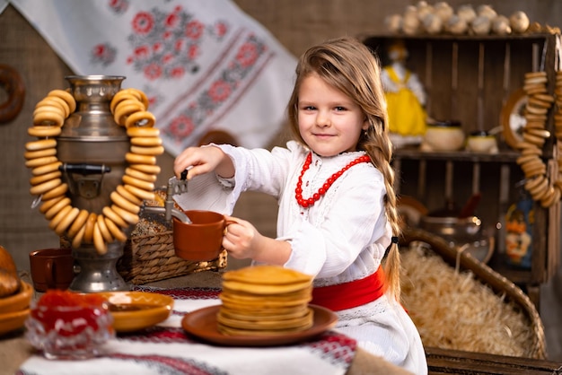 Piccola ragazza caucasica in costume popolare che versa il tè dal samovar mentre celebra Maslenitsa