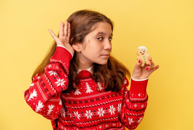 Piccola ragazza caucasica in possesso di biscotti di Natale isolati su sfondo giallo cercando di ascoltare un pettegolezzo.