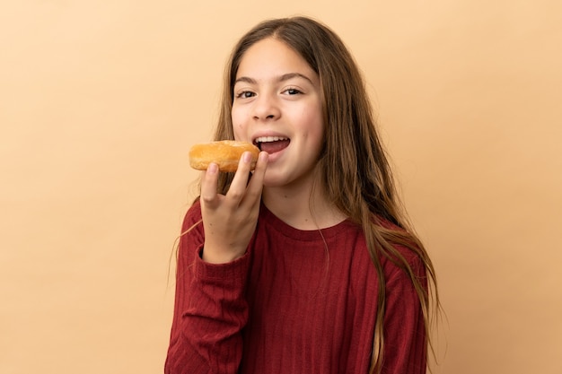 Piccola ragazza caucasica isolata su fondo beige che mangia una ciambella