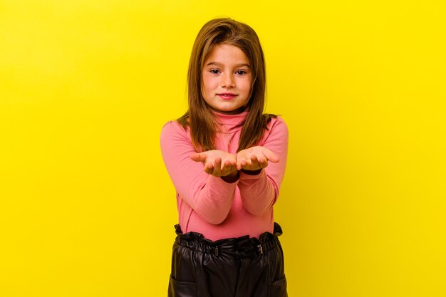 Piccola ragazza caucasica isolata su giallo che tiene qualcosa con le palme, offrendo alla macchina fotografica.