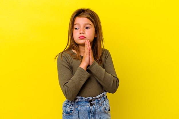 Piccola ragazza caucasica isolata su giallo pregando, mostrando devozione, persona religiosa in cerca di ispirazione divina.