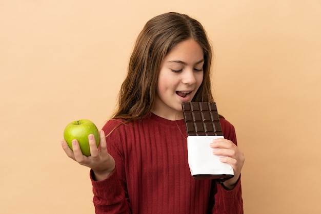 Piccola ragazza caucasica isolata su sfondo beige che prende una tavoletta di cioccolato in una mano e una mela nell'altra