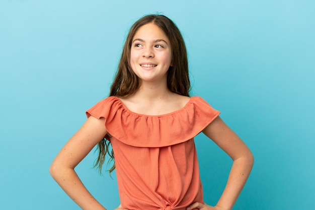 Piccola ragazza caucasica isolata su sfondo blu in posa con le braccia all'anca e sorridente