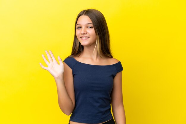 Piccola ragazza caucasica isolata su sfondo giallo che saluta con la mano con espressione felice