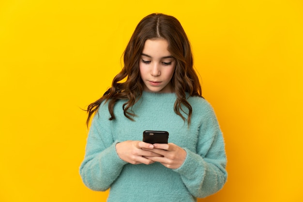 Piccola ragazza caucasica isolata su sfondo giallo utilizzando il telefono cellulare