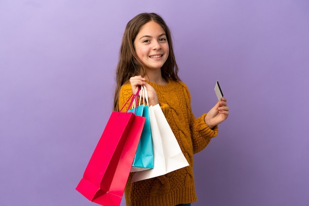 Piccola ragazza caucasica isolata su sfondo viola con borse della spesa e una carta di credito